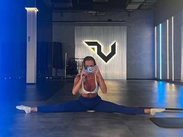 JessicaAubrey is doing a full front split in a gym, wearing a white sports bra, blue leggings, and white socks, with her tits visible and taking a selfie.