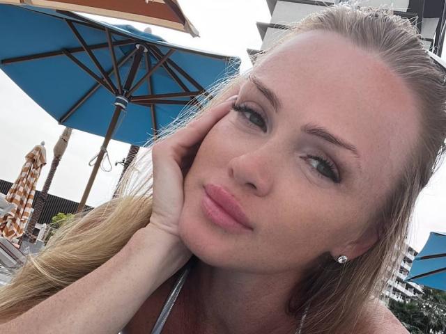 JessicaAubrey with blonde hair, pink lips, green eyes, and diamond earrings, touching her face with a slight smile, wearing a halter top, under blue umbrellas.