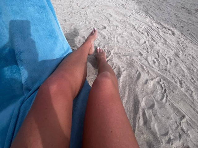 JessicaAubrey's tanned, sunburned legs with sand on her feet, lying on a blue towel at a sandy beach, her shadow on the towel.