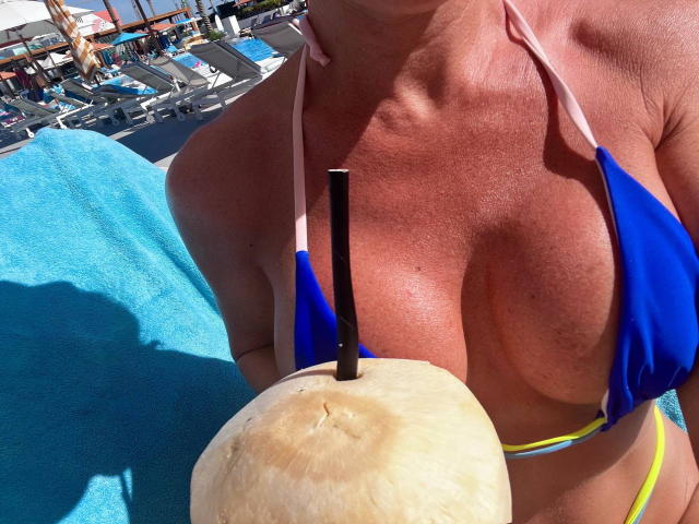 JessicaAubrey's sun-kissed tits in a blue bikini, holding a coconut with a straw, sitting by a bright blue pool. Her yellow bikini bottom is visible.