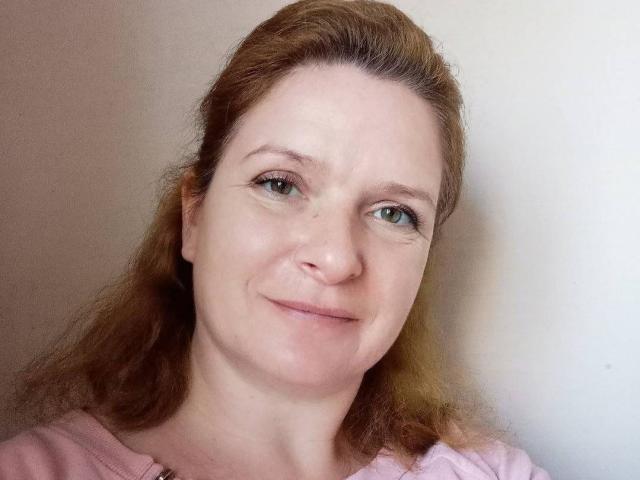 HelenEvansy has light skin, green eyes, and brown wavy hair, wearing a pink shirt, with a slight smile and subtle makeup. Her nose is slightly upturned.