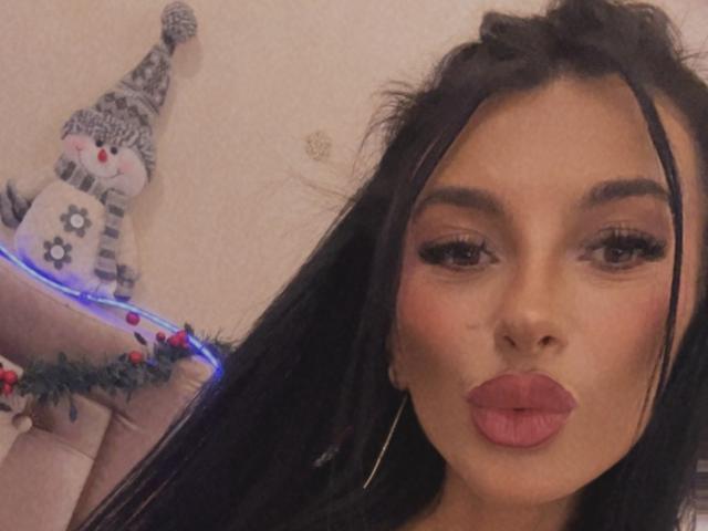 KinkyAmelie with pouty pink lips, long black hair, and thick eyelashes, holding a snowman figurine with blue lights and red ornaments. Boobs partially visible in the background.