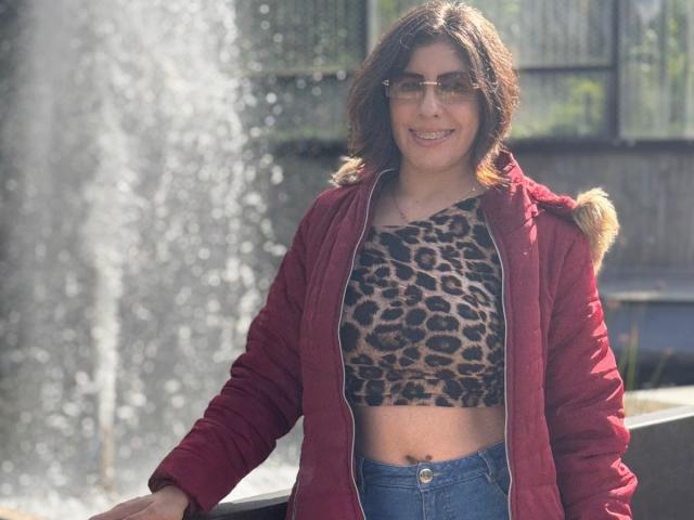 AnastasiaEvansX with medium brown curly hair, glasses, red jacket, and leopard print crop top, standing by a water fountain, smiling with blue jeans and her ass slightly exposed.