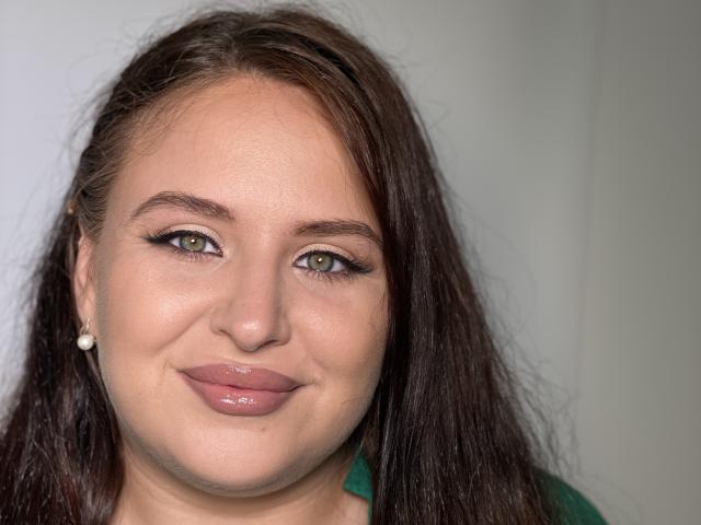 CrystalFaye has green eyes, full pink lips, and dark brown, wavy hair. She's wearing small pearl earrings and a green top. Her face is slightly flushed.