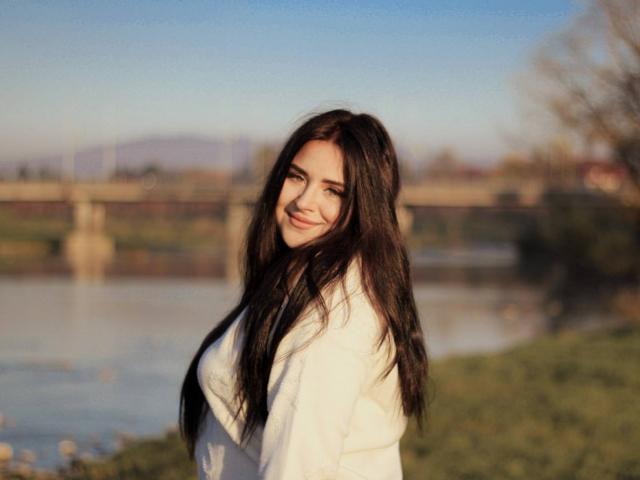 CrystalFaye with long dark hair, smiling, wearing a white coat, standing by a calm river, sunlit, soft focus, blue sky, green grass, and a bridge in the background.