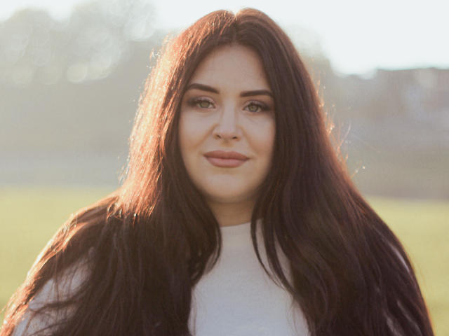 CrystalFaye with long, dark brown hair, light skin, green eyes, and a subtle smile, wearing a white top, sunlight highlighting her hair.
