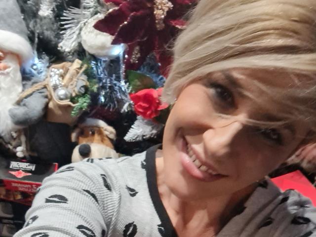InanAIshtars met blond haar, lachend, die een grijze trui met zwarte patronen dragen, met zichtbare tanden, in een kerstthema-omgeving met decoraties, waaronder een kerstman en een honden speelgoed..