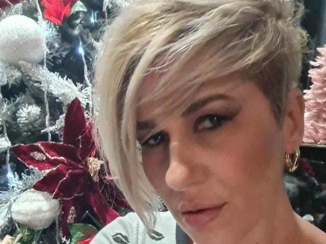 Vrouwen met kort blond haar, volle lippen en gouden oorbellen, met een subtiele grijns. Kerstboom met rode kerststerren en witte versieringen op de achtergrond..