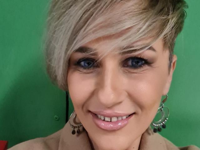 InanAIshtars tiene el cabello corto y rubio, ojos azules y unos labios rosados brillantes, con aros grandes, y está sonriendo a la cámara. No se ven los senos..