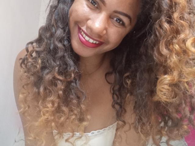 LayahCoquine with curly brown and blonde hair, smiling with pink lipstick, showing her white strapless top and bare shoulders, looking cute and sexy.