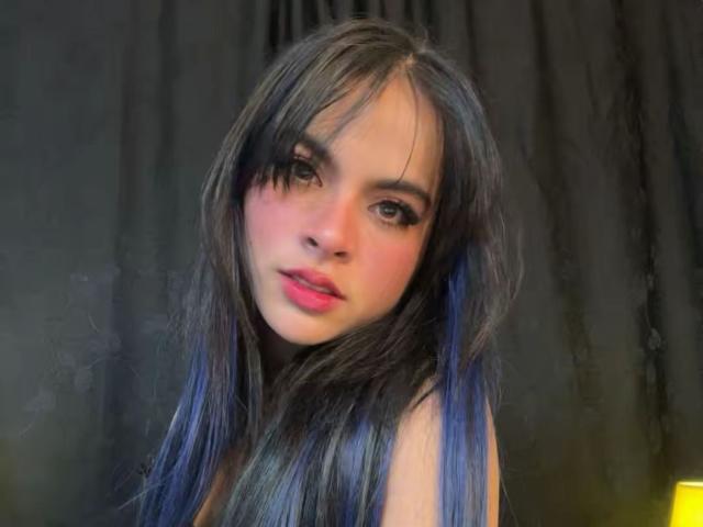FireCoupleHot with long, dark, blue-tinted hair, pink lips, and brown eyes, looking slightly to the side with a soft, innocent expression. Dark background, blurry.