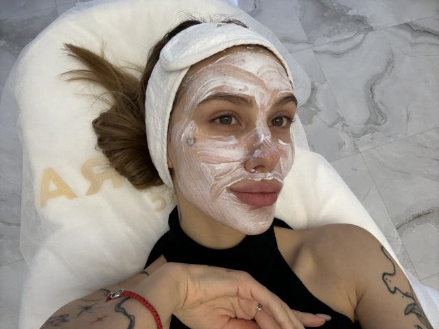 SensualMila lying on a white pillow with white towel on head, face covered in thick white mask, black sleeveless top, tattoos on arms, marble floor, red bracelet.