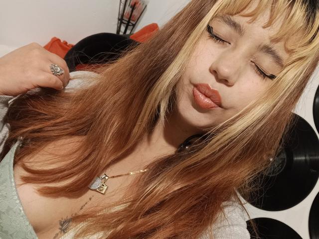 BelleVivienneHt met gesloten ogen, volgeblazen lippen en blond haar, draagt een groene top, een gouden ketting en een ring, en houdt haar haar vast. Haar borsten zijn licht zichtbaar..