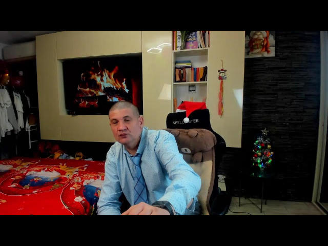 CrisSalon, bald with a light blue shirt, leans on a chair with "DIPPER" on it, in a room with a red bedspread, a fire TV screen, and a Christmas tree.