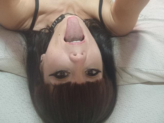OrianaLaFrancaise's face upside down, tongue out, looking at the camera, black hair, dark eyes, wearing a black strap, white pillow under her head.
