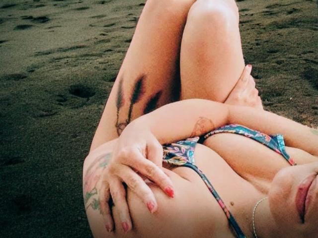 RosannaBlue's tits are small in a colorful bikini, lying on dark sand with crossed legs, tattoo on right thigh, red nail polish, and hands covering her chest.