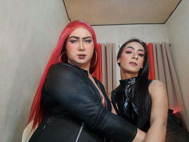 TwoAsianShemales with red hair, large boobs, black leather jacket, and hoop earrings, standing next to TwoAsianShemales with black hair, shiny black top, and pink eyeliner.