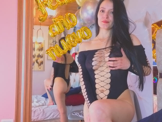 AriaBonnet, avec de longs cheveux noirs, portant une combinaison noire en filet, sans manches, qui expose ses seins et ses parties intimes. Des ballons dorés en forme de "20" sont visibles en arrière-plan. Un miroir réfléchissant..