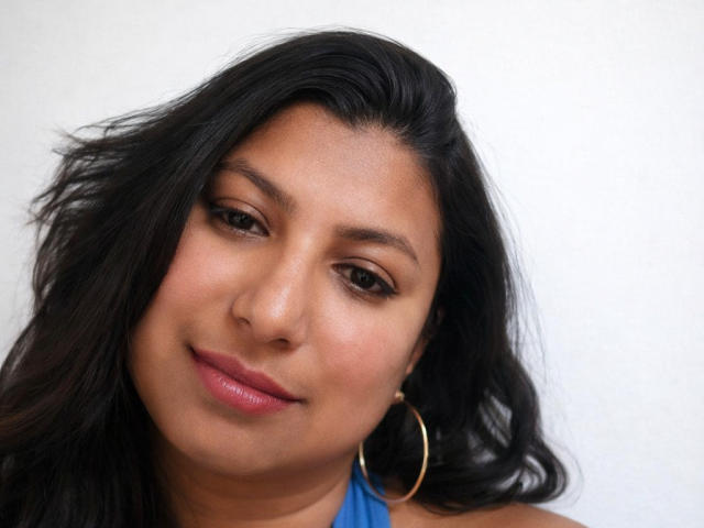 LunaExotica has medium brown skin, black wavy hair, hoop earrings, and pink lipstick. She's smiling slightly with brown eyes looking forward. Her blue top is visible.