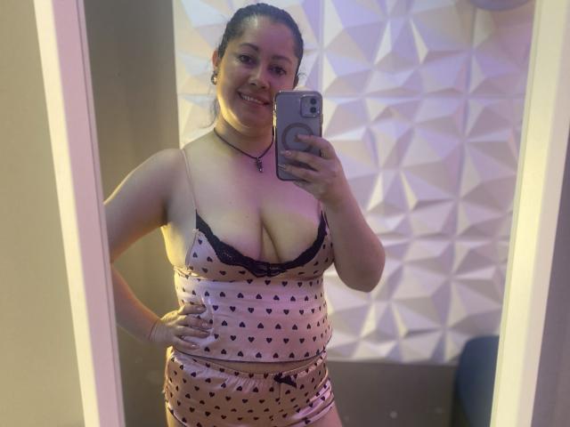 MarianaSmitt with dark braided hair, smiling, wearing a pink camisole with black lace and heart patterns, matching pink shorts, holding a phone, showing her big tits.