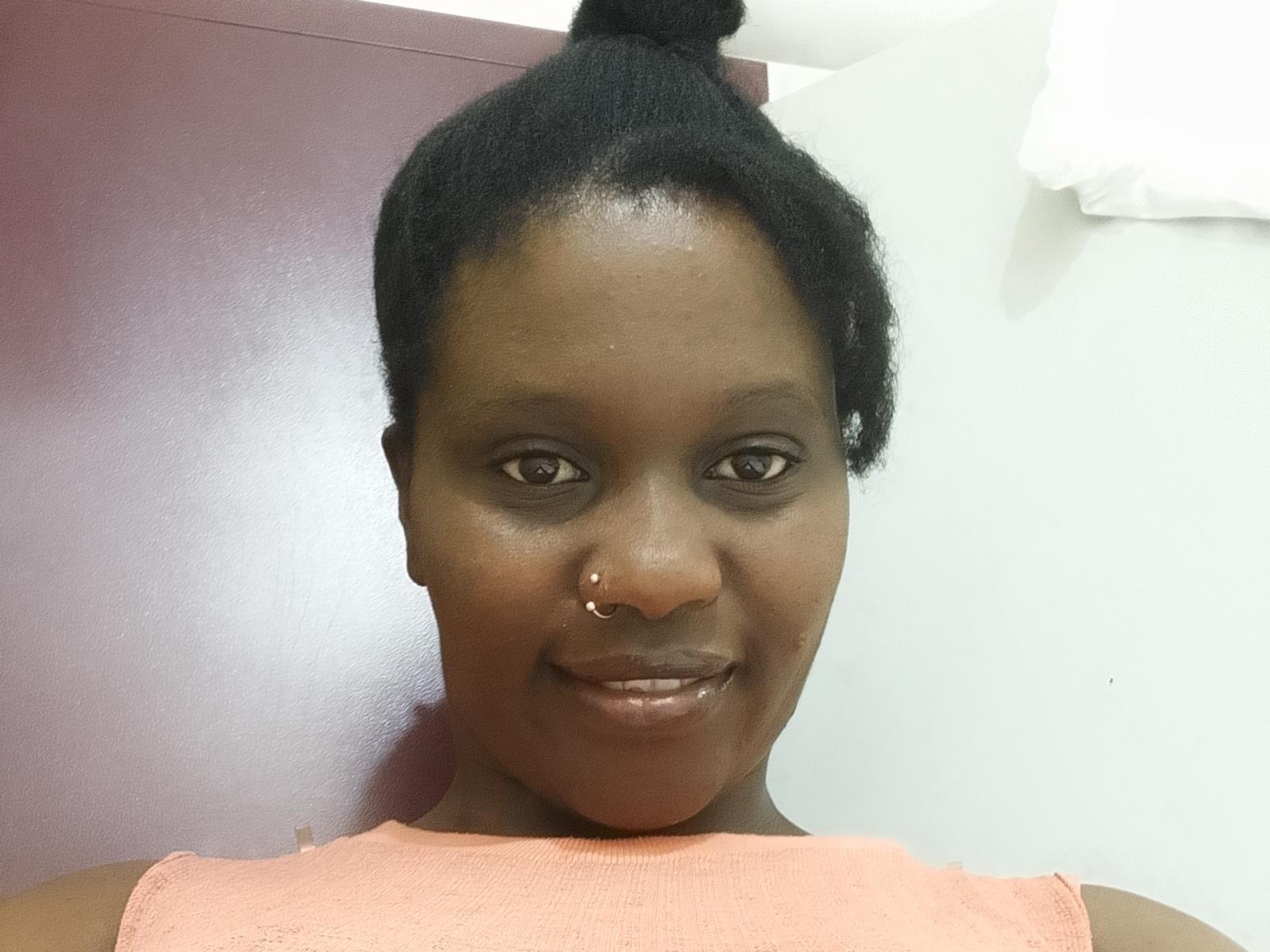 Hustion tem a pele escura, cabelo preto preso em um coque, um piercing no nariz e um sorriso. Ela está vestindo uma blusa rosa sem mangas, que mostra seu rosto brilhante e pele lisa..