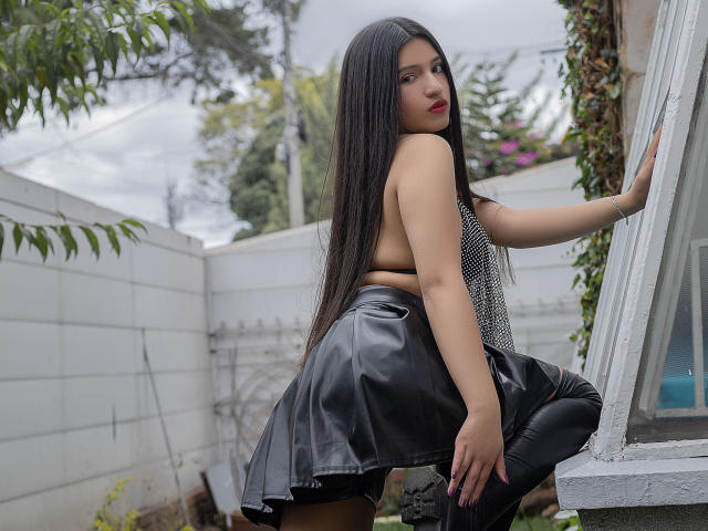 LaureneLove with long black hair, red lipstick, and a shiny black leather skirt, squatting, showing her ass, leaning on a window, wearing a sparkly top, and black boots.