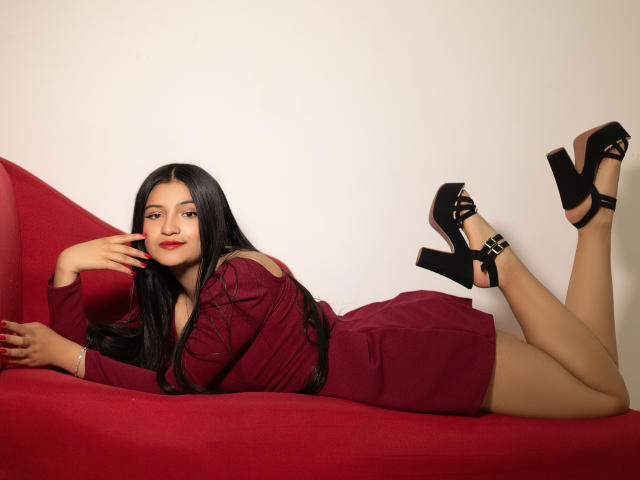 LaureneLove lies on a red chaise, legs bent at the knees with feet up, wearing a maroon dress and black high-heeled sandals with buckles. Her long black hair is straight, and she has red lipstick. She rests her right hand near her face, which has a subtle smile. The maroon dress has long sleeves and a belt. The image has a soft, even lighting with a clean, white background. Her skin is light brown. The chaise is positioned horizontally.