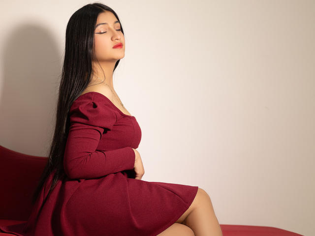 LaureneLove with long black hair, closed eyes, and red lipstick, wearing a red dress, sitting on a red chair, showing her ass and tits, looking relaxed and sexy.
