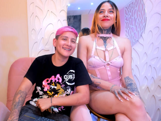 HadeDanny with pink hair, black shirt, and tattoos sits next to HadeDanny with blonde hair, pink lingerie, and tattoos, showing tits and a tattooed neck. Smiling, they're on a pink chair.