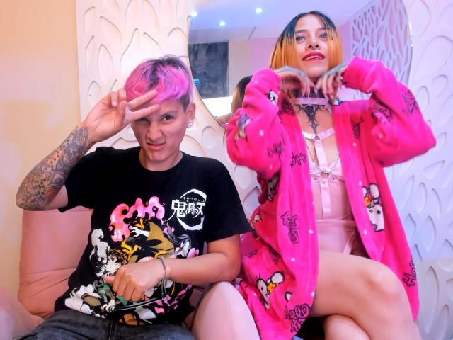 HadeDanny with pink hair, tattoos, and black t-shirt, making a peace sign. HadeDanny in pink robe, lingerie, and makeup, flashing heart. Both sitting on pink chair.