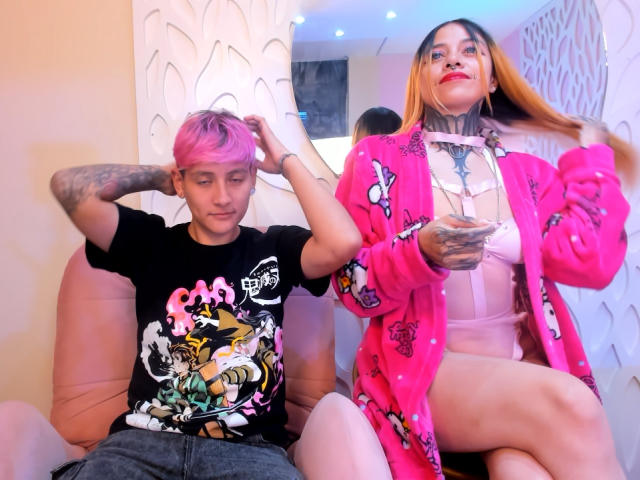 HadeDanny with pink hair, tattoos, and black shirt sits on a pink chair. HadeDanny with long brown hair, pink cardigan, and pink lingerie stands, showing tattoos and red lips.