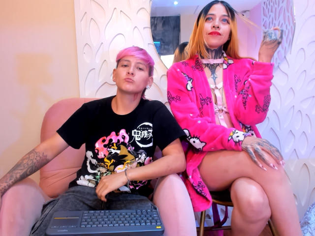 HadeDanny mit rosa Haaren, schwarzem Hemd und Tätowierungen sitzt auf einem rosa Stuhl, ein Laptop auf dem Schoß. HadeDanny mit orange-braunen Haaren, einem rosa Schmetterlingsbademantel und Tätowierungen sitzt auf einem Stuhl..