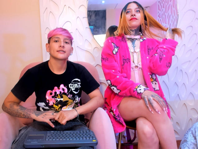 HadeDanny with pink hair, black shirt, and tattoos sits with HadeDanny in pink bunny robe, tattoos, and red lipstick, holding hair, both with laptops, in a pink chair.