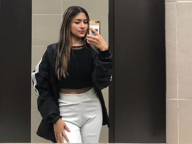 AfroditaWild takes a mirror selfie. She has long, straight brown hair, fair skin, and wears a black, long-sleeve crop top with a sheer neckline, white high-waist pants, and a black jacket with white stripes on the sleeves. She holds a gold-colored phone in her right hand, looking to the side with a slight smile. The image has a casual, modern style with a focus on her outfit and reflection.