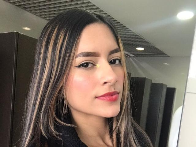 AfroditaWild with long brown hair, black eyeliner, pink lipstick, and a slight smile, wearing a black top, in a modern room. Her nose is small and her skin is fair.