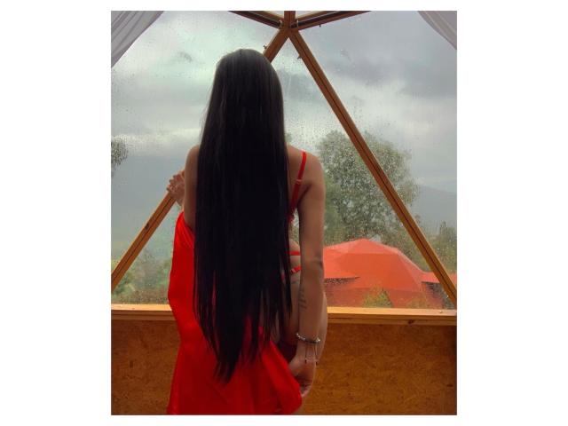 ThaliaCollins with long black hair, red dress, and silver bracelet, standing in front of a wooden framed window, looking outside at a cloudy, wet day. Boobs visible from behind.