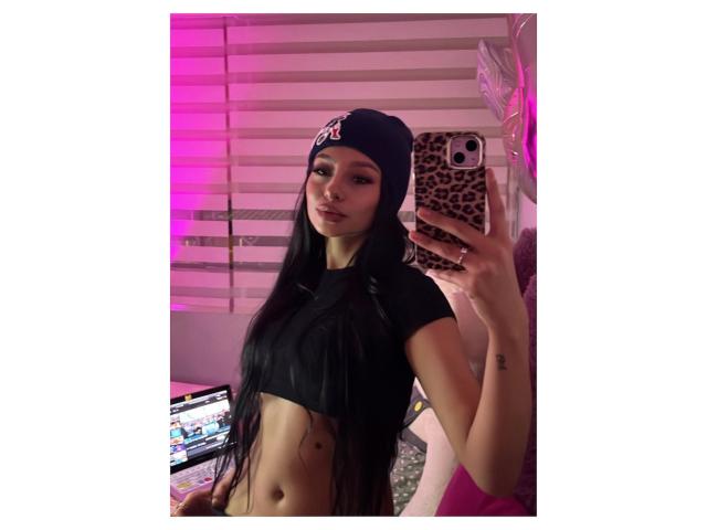 ThaliaCollins with long black hair, wearing a black crop top and leopard print beanie, taking a mirror selfie with a leopard phone case, pink lighting in the background.