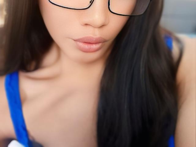 AlluringAmelia with dark straight hair, glasses, and pouty pink lips, wearing a blue strap top, showing her cleavage and smooth tan skin.