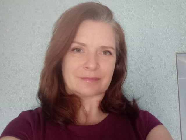 HelenEvansy with brown hair, green eyes, and a slight smile, wearing a purple shirt, in a selfie, showing her face and upper body. Her ass is not visible.
