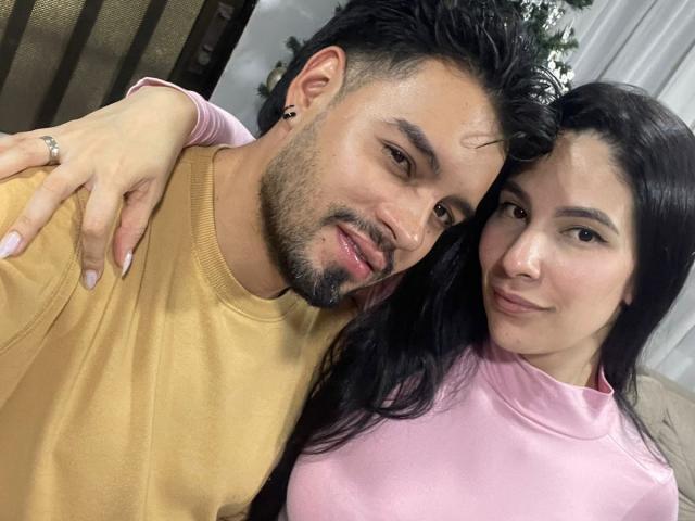 AriannaAndDave, a cute mixed couple, Arianna with long black hair in a pink shirt, Dave with a beard in a yellow shirt, smiling and touching shoulders.