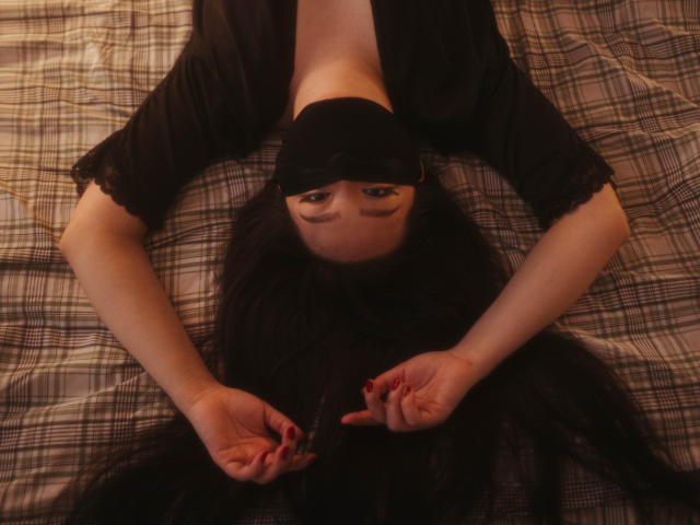 BeloverNana is lying on her back with arms above her head, black hair spread out, wearing a black top, holding her hair with red nails, looking up.