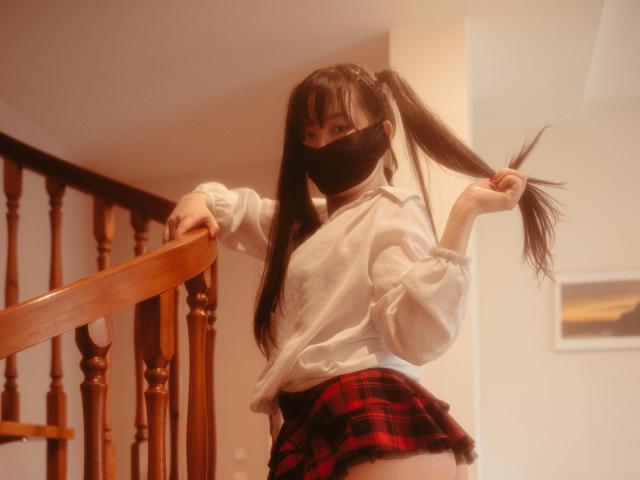 BeloverNana with pigtails, wearing a white blouse, red plaid skirt, and black face mask, leaning on a wooden staircase, showing her ass, in a warm, soft-focused shot.