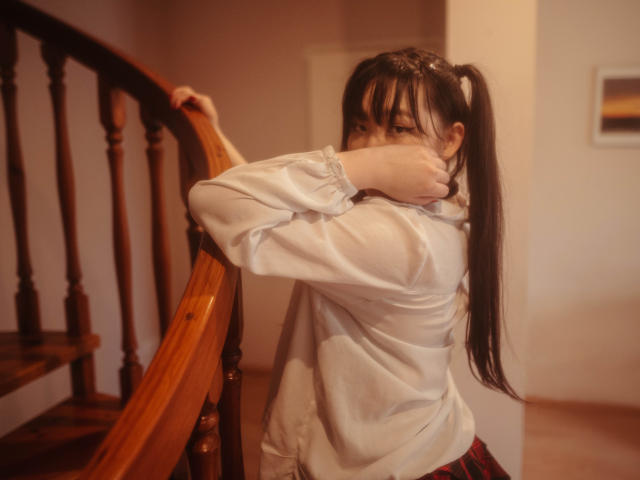BeloverNana stands on a wooden staircase, arms crossed and face partially hidden. She wears a white, oversized shirt and a red plaid skirt. Her long, dark hair is tied in a high ponytail. The wooden staircase has a polished, warm brown finish. The light is soft and warm, casting a gentle glow on BeloverNana's face and the staircase. The composition emphasizes her modest posture and the contrast between the white shirt and the wooden staircase. The image has a slightly blurred, vintage filter effect.