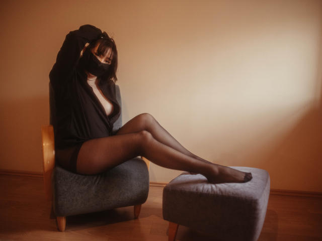 BeloverNana with long legs, black sheer stockings, and black top, sitting on a grey chair, legs on matching ottoman, arms covering face, wooden floor, dim lighting.