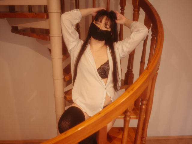 BeloverNana with long black hair, wearing an open white shirt, black lace bra, and black mask, has hands in hair, black thigh-highs, sitting on wooden spiral staircase.