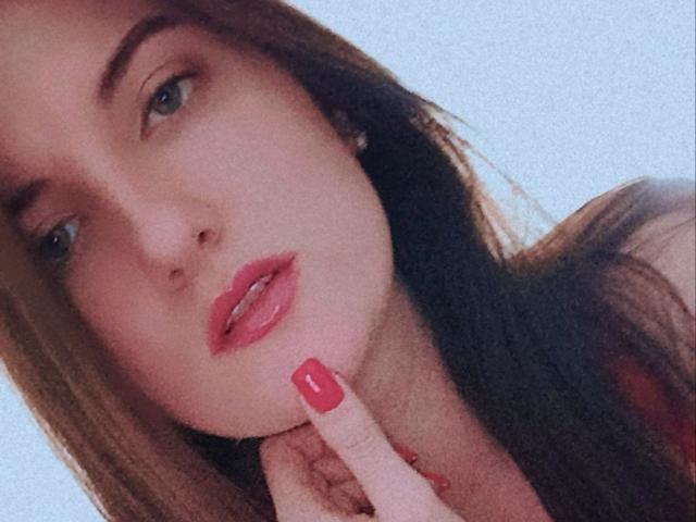 StephieRouse's face, with red lips, blue eyes, and brown hair, touching her chin with a finger that has red-painted nails. She looks slightly seductive and relaxed.