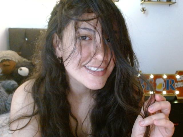 MargaritaDudole with messy black hair, smiling, holding a lock of hair, showing her white teeth, topless, with small tits, in a bedroom, wearing a playful expression.