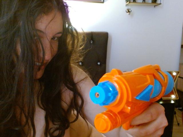 MargaritaDudole with long, wet brown hair, holding an orange and blue water gun, her bare tits partially visible, in a dimly lit room.
