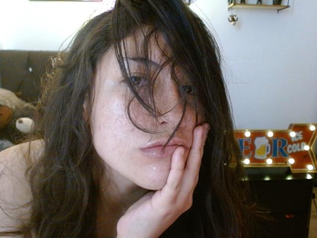 MargaritaDudole with messy black hair, freckles, and glossy lips, touching her cheek with her hand, looking slightly to the side. Her tits are partially visible.