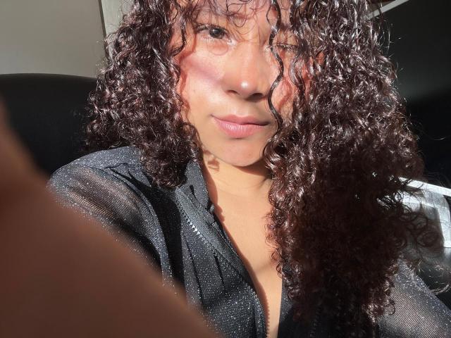 SofiaPassionne with curly brown hair, glowing tan skin, and a subtle smile, wearing a black sheer top, sunlight highlighting her face. Boobs partially visible under the top.